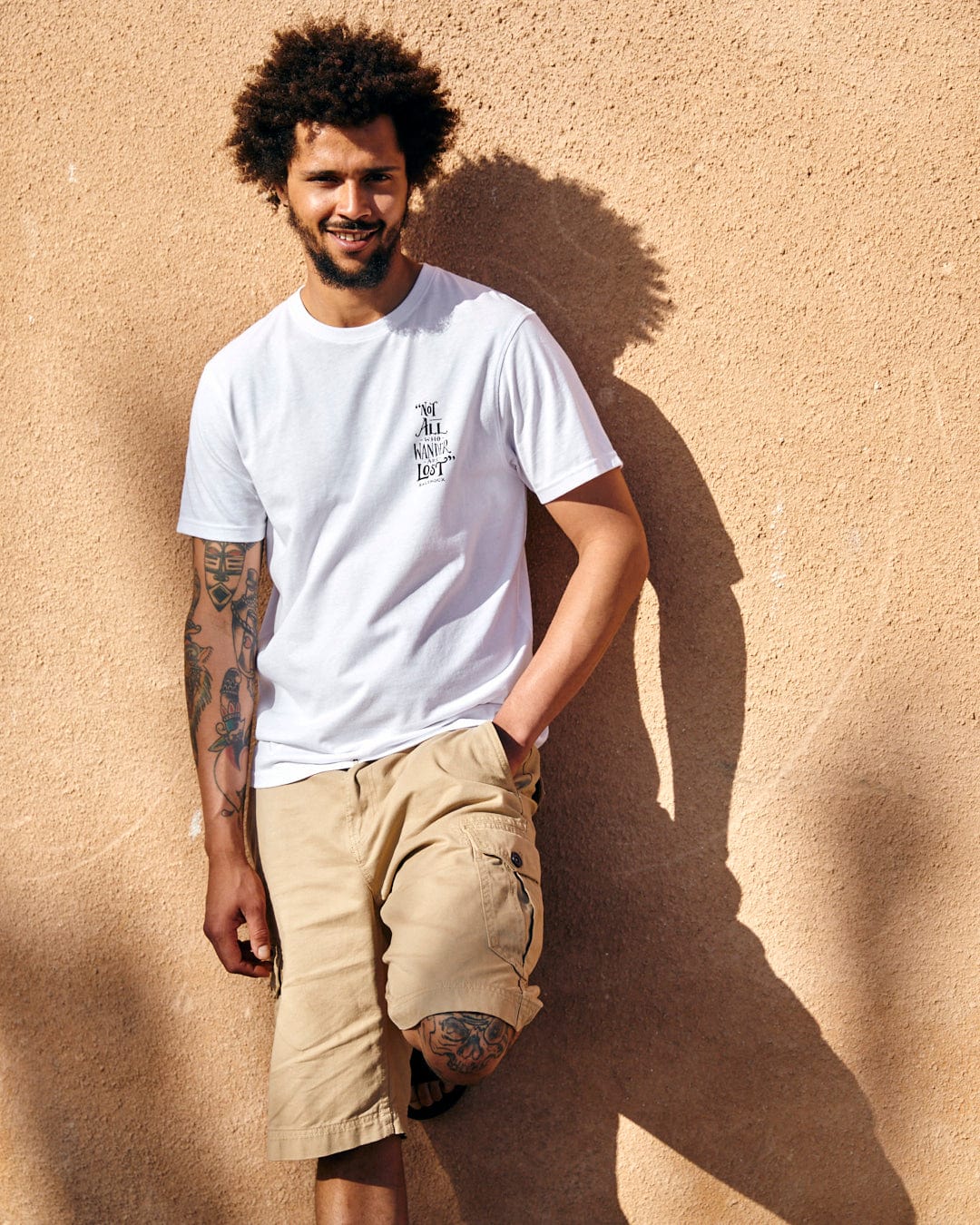 Young man with curly hair and tattoos smiling while leaning against a sunlit beige wall, wearing a Saltrock Lost Ships Short Sleeve T-Shirt in White and tan shorts.