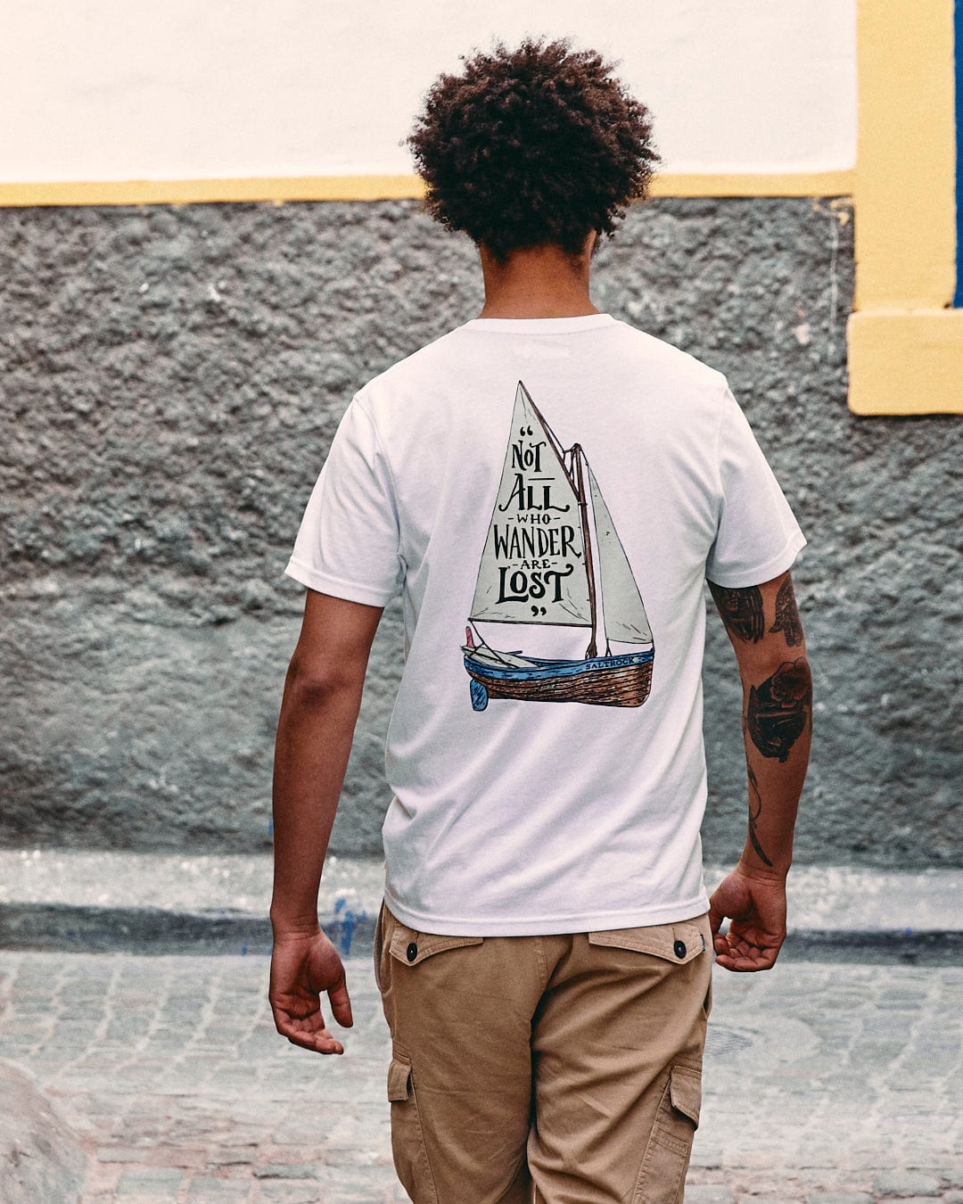 A person standing in an alley, facing away, wearing a Lost Ships Short Sleeve T-Shirt in White by Saltrock, paired with beige shorts.