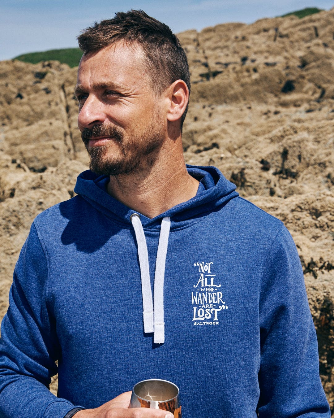 A man wears the Saltrock Lost Ships Men's Pop Hoodie in Blue Marl, featuring a "Not all who wander are lost" design, a drawcord hood, and a kangaroo pocket. He stands outdoors with a metal cup against rocky terrain. Crafted from recycled polyester for eco-friendly adventurers.