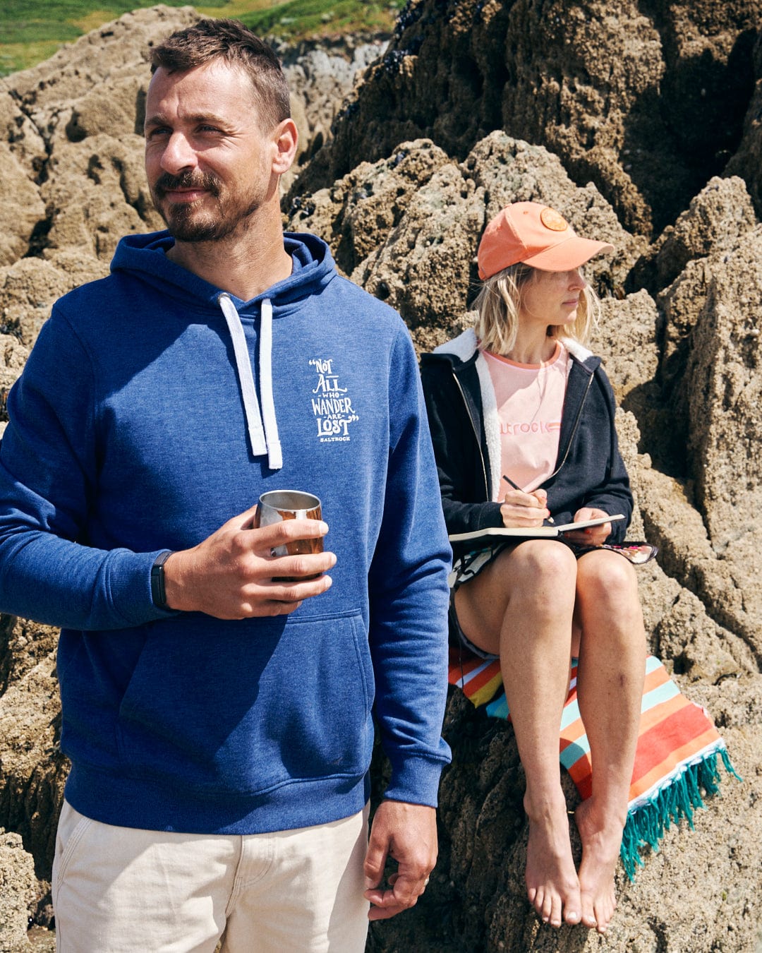 A man in a Lost Ships - Mens Pop Hoodie by Saltrock, featuring a drawcord hood and kangaroo pocket, holds a drink while standing on rocks. Nearby, a woman in a pink cap sits on a striped towel with a book, enjoying the serene setting.