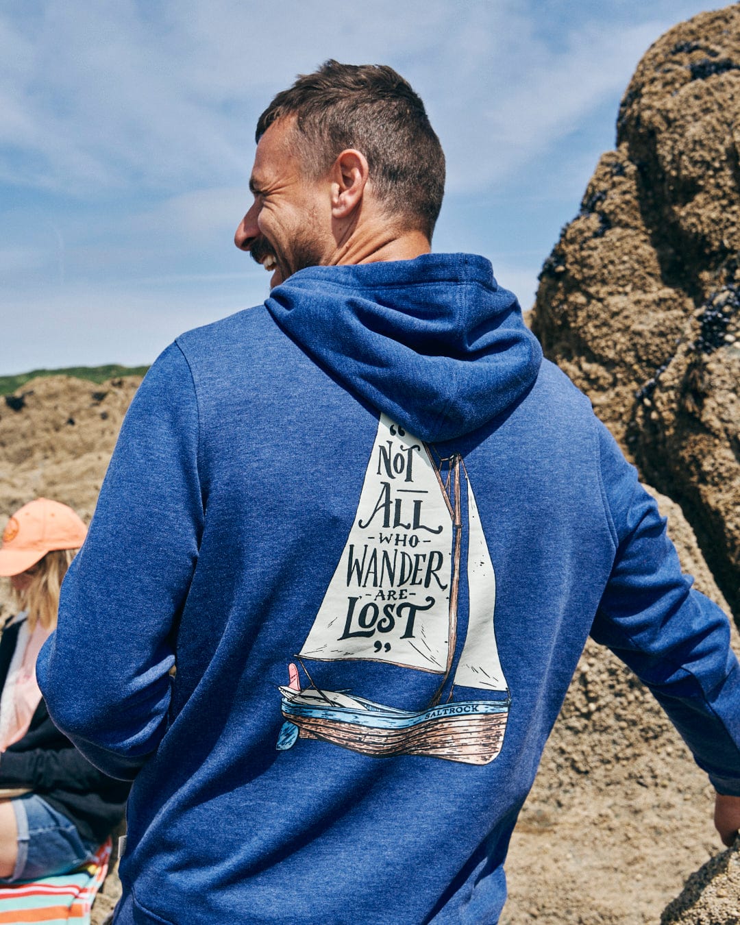 A man stands near rocky terrain wearing the Saltrock Lost Ships Men's Pop Hoodie in Blue Marl, made from recycled polyester. It features a sailboat design with "Not all who wander are lost," and includes a kangaroo pocket and drawcord hood for comfort.