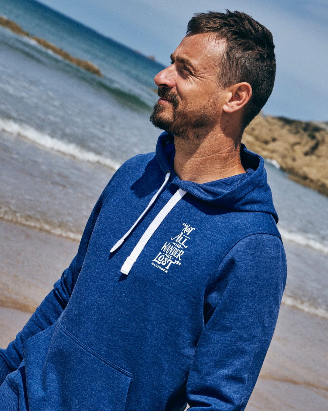 A man wearing the Saltrock Lost Ships Men's Pop Hoodie in Blue Marl, made from recycled polyester, stands on a beach gazing at the ocean with sky and rocks behind him, his hands warmly nestled in its kangaroo pocket.