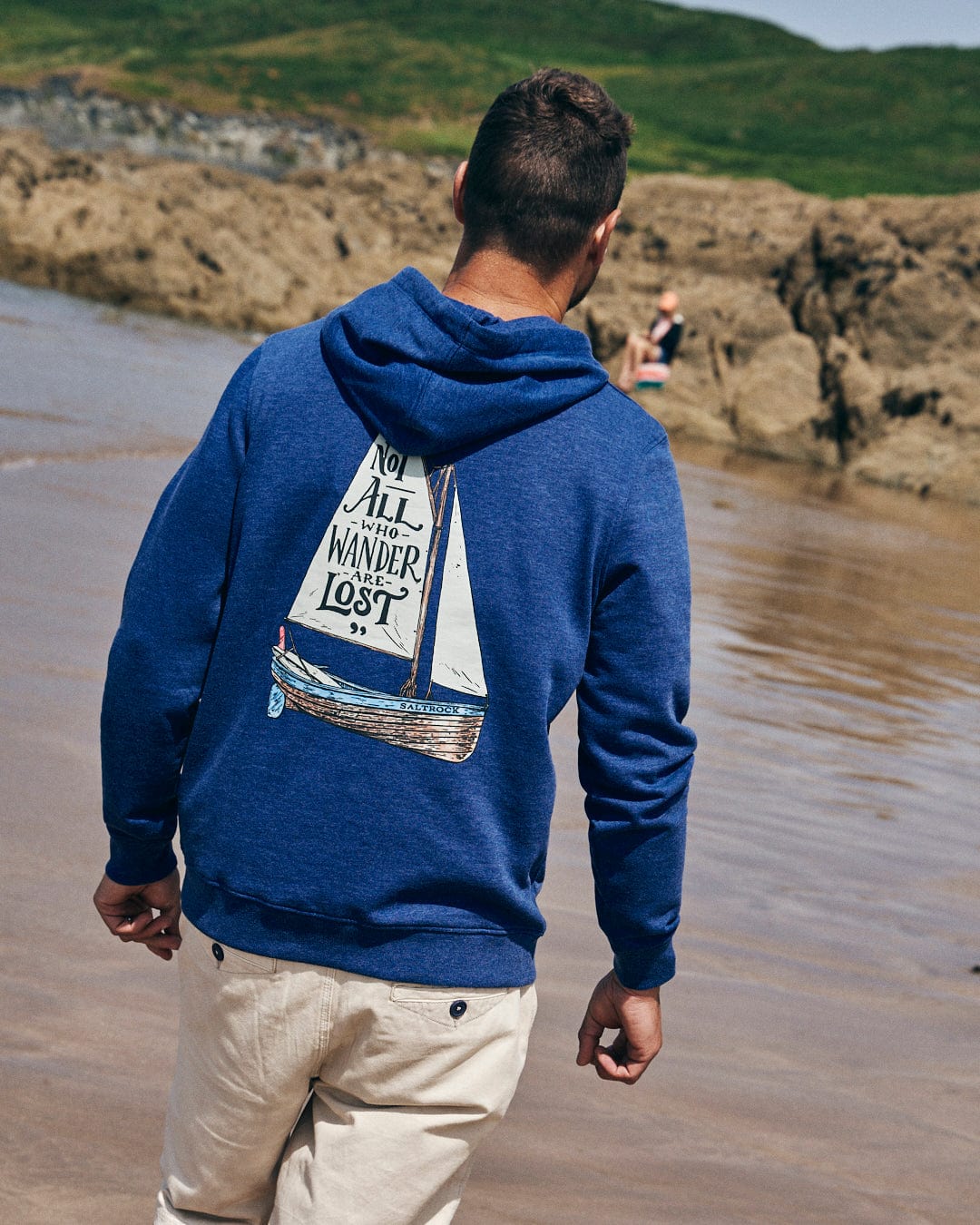 Standing on a beach, a person wears the Saltrock Lost Ships Men's Pop Hoodie in Blue Marl, featuring a sailboat graphic with "Not All Who Wander Are Lost." This cozy hoodie, crafted from recycled polyester, includes a drawcord hood and kangaroo pocket. Rocks and grass fill the backdrop.