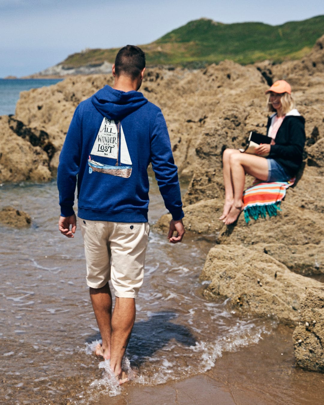 A man in the Saltrock Lost Ships Men's Pop Hoodie - Blue Marl takes a walk on the rocky beach, near the water's edge, as a woman wearing a cap unwinds on her towel with a book.