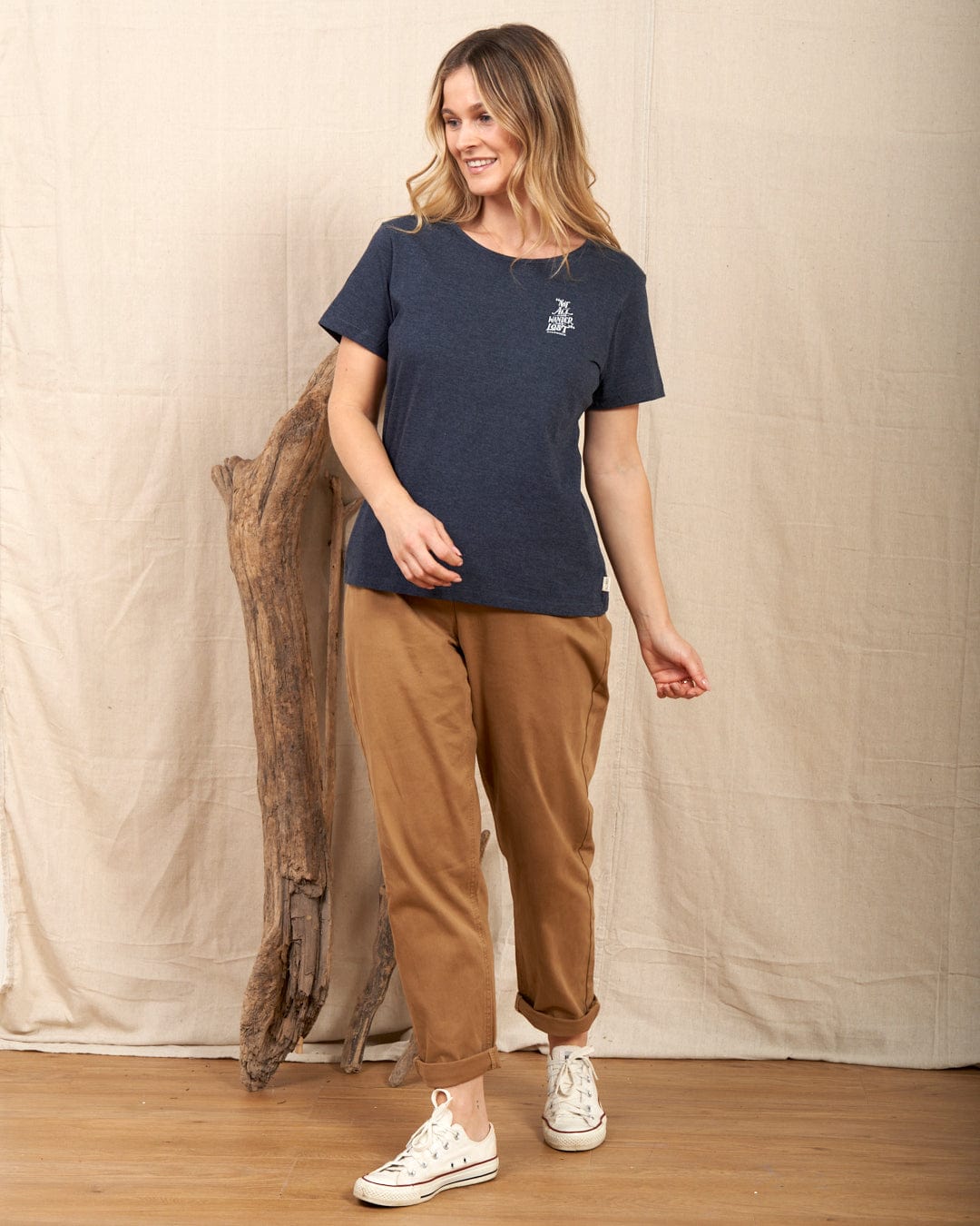 A woman in a Saltrock Lost Ships navy T-shirt and brown pants smiles indoors, standing by a wooden log against a beige backdrop. She sports white sneakers, and her machine-washable outfit is ideal for everyday adventures.