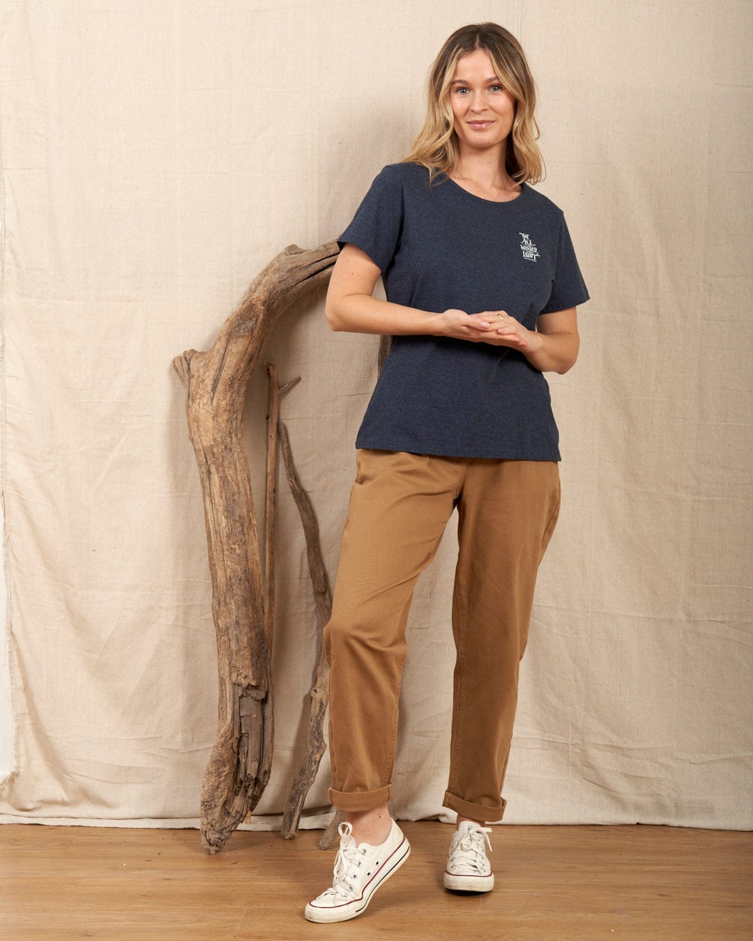 A person wearing a Saltrock "Lost Ships" women's T-shirt in dark blue and brown pants stands indoors beside a driftwood piece, expression neutral, on a wooden floor with a fabric backdrop.