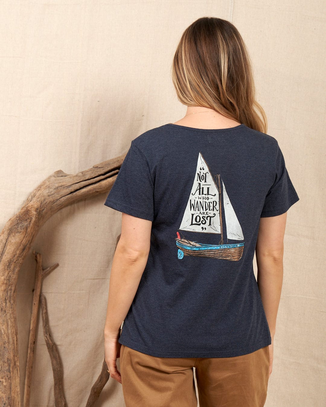 A woman with long hair wears the Saltrock "Lost Ships" women's T-shirt in blue, featuring a sailboat and "Not All Who Wander Are Lost" on the back. She stands before a beige fabric backdrop decorated with driftwood. Made of 100% cotton.