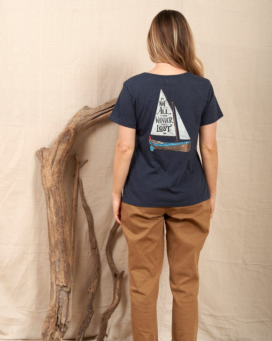 A woman with long hair faces away, wearing the Saltrock Lost Ships Women's T-Shirt in blue, crafted from 100% cotton and featuring a sailboat design and "Not all who wander are lost" text, standing by driftwood embodying the spirit of exploration.