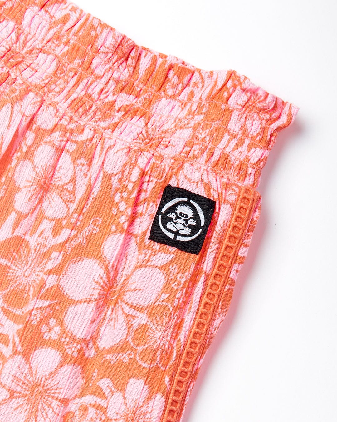 Close-up of Lottie Hibiscus - Kids Trousers - Orange/Pink fabric with a shirred waistband and a Saltrock logo patch.