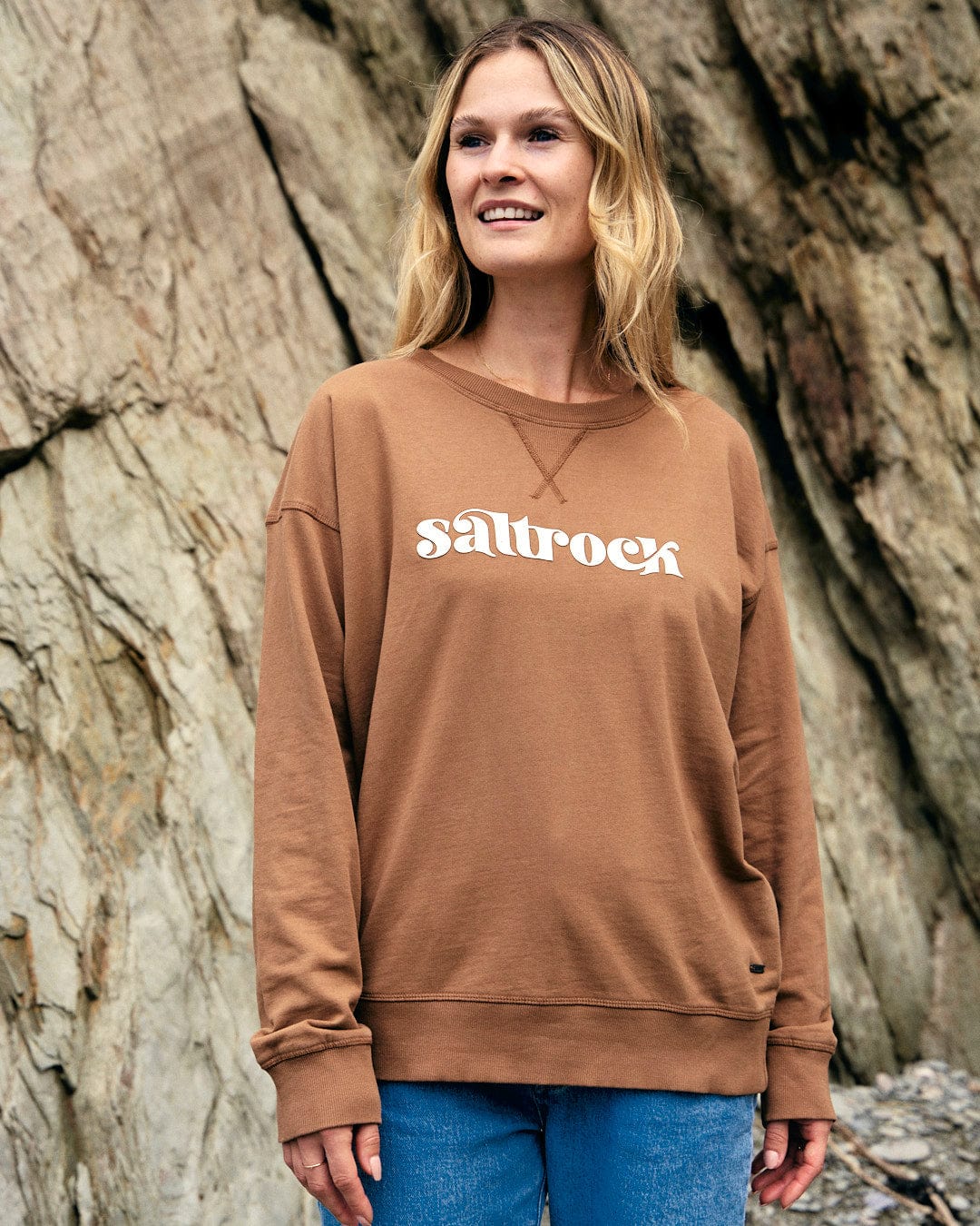 A woman wearing a brown Saltrock Louisa - Womens Crew Sweat - Brown sweatshirt standing in front of a rock.