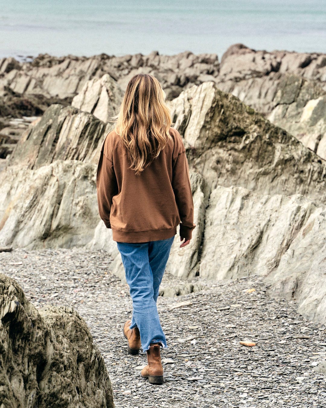 Louisa walking on a rocky path near the ocean, wearing a Saltrock Louisa - Womens Crew Sweat - Brown cotton outfit.