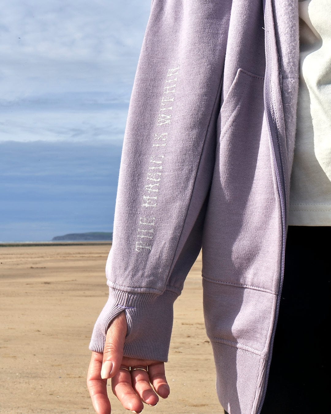 A woman wearing a Saltrock Magic Within - Women's Lilac Zip Hoodie with a floral skeleton design on the beach.