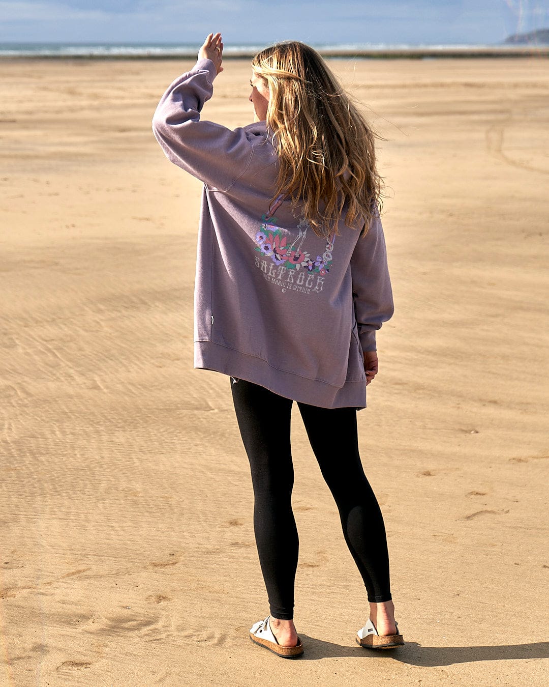 A woman standing on a beach wearing a Lilac Saltrock Magic Within - Womens Zip Hoodie with a floral skeleton design.