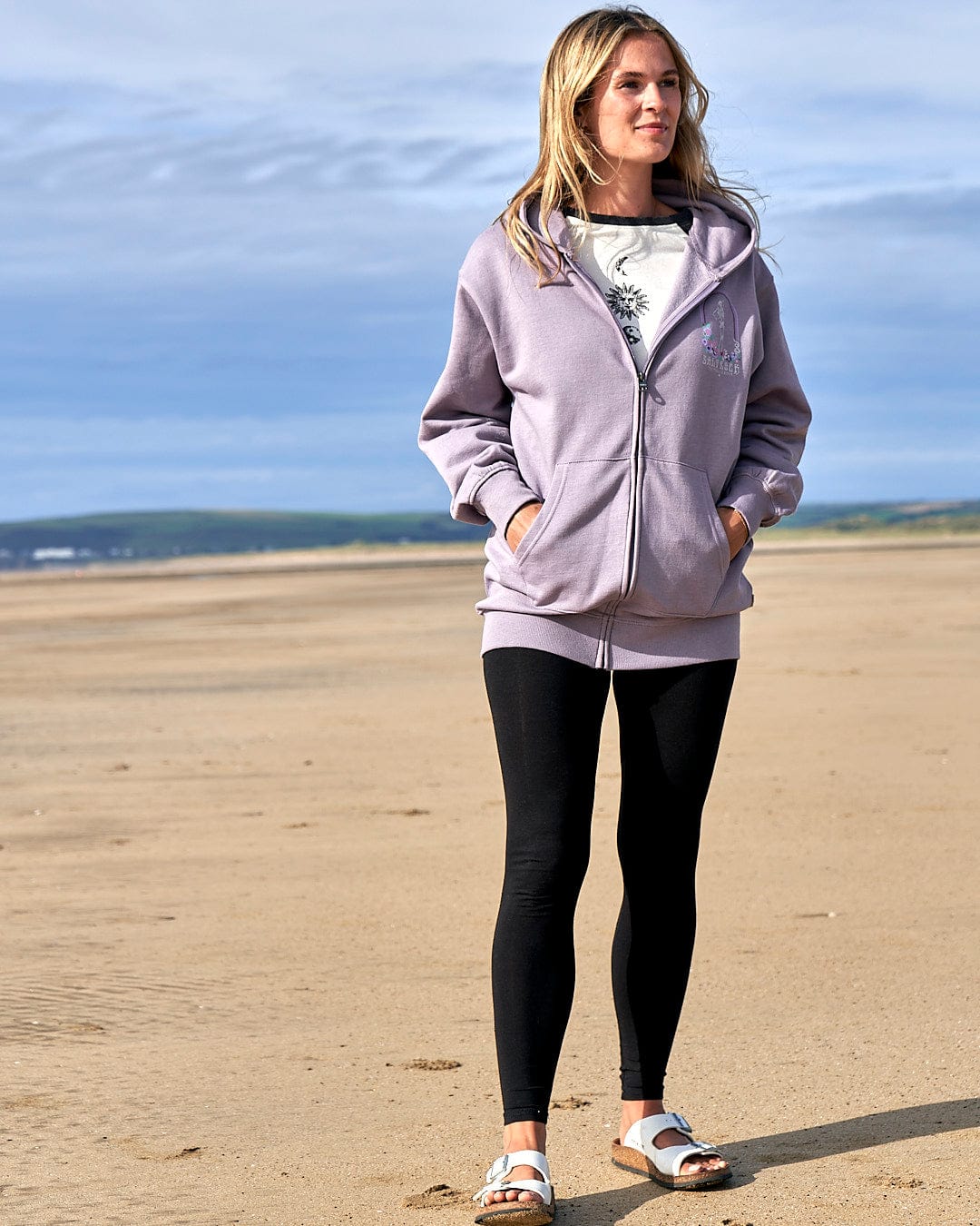 A woman standing on a beach wearing a Saltrock Lilac Magic Within - Womens Zip Hoodie and black leggings, featuring a floral skeleton design.