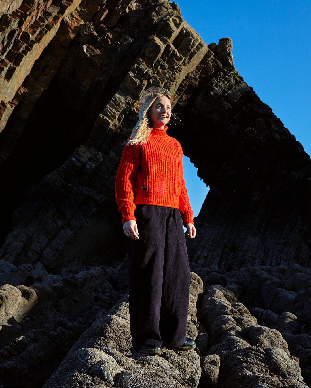 A woman in a red sweater standing on rocks, wearing a Saltrock Mandy - Womens Funnel Neck Jumper - Red.