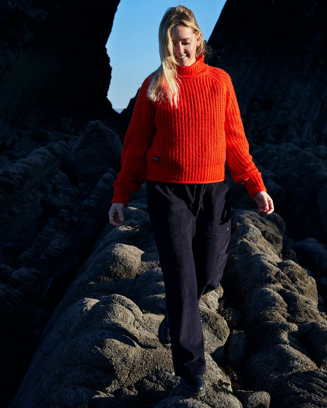 A woman in a Saltrock Mandy - Womens Funnel Neck Jumper - Red walking on rocks.