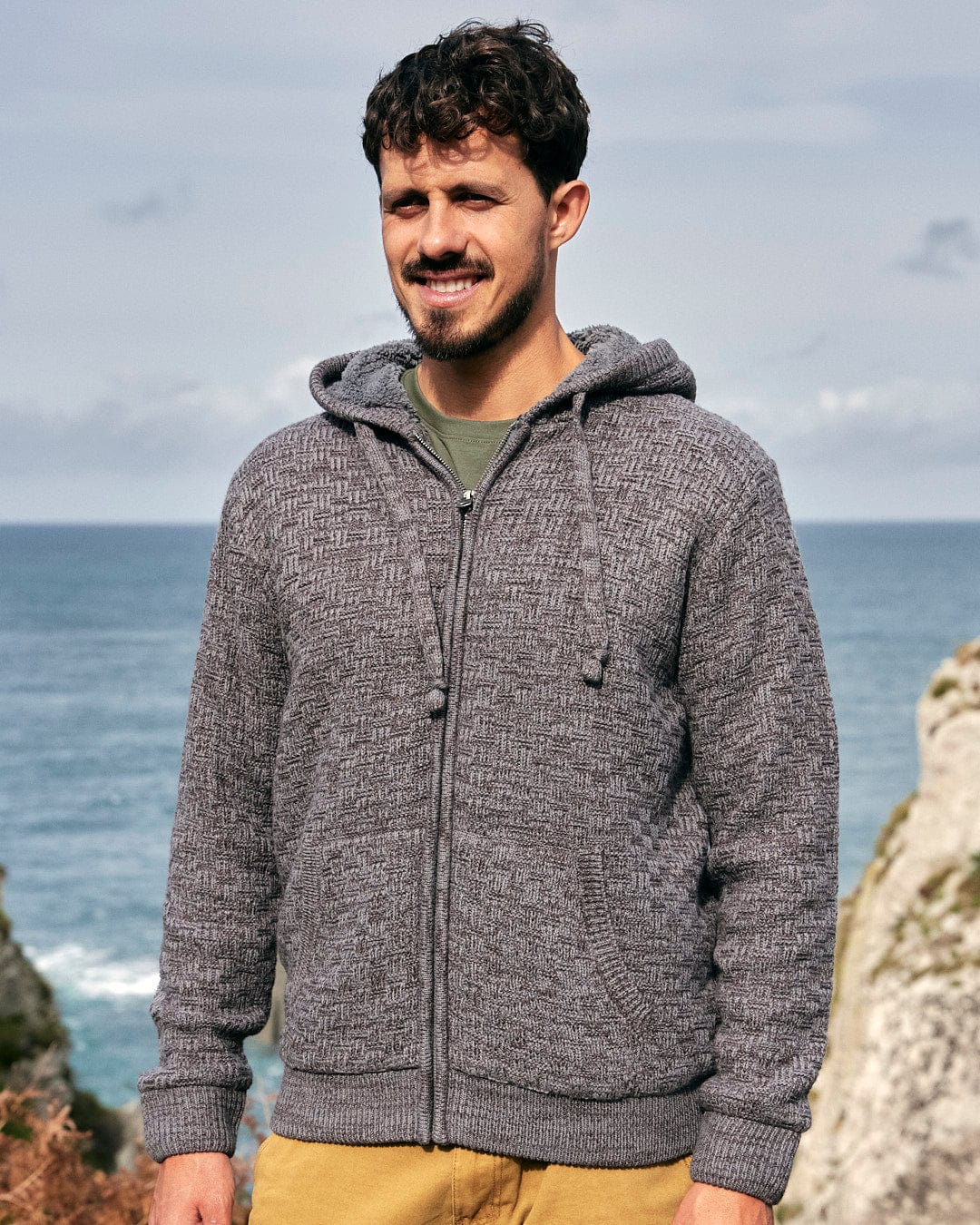 A man modeling the Saltrock branding, wearing a Markus - Mens Borg Lined Knit Hoodie - Dark Grey for warmth, stands next to the ocean.