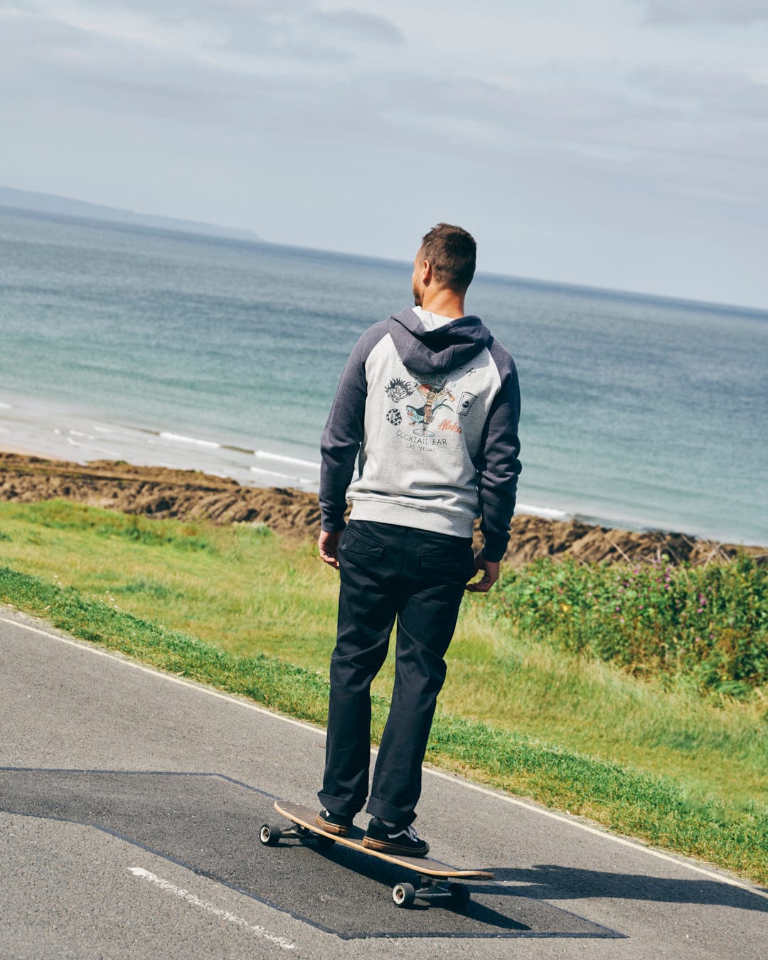 Wearing Saltrock's Meltan Mens Straight Leg Twill Trouser in Dark Grey, a person skateboards along a paved coastal road with green grass and ocean scenery in the background.