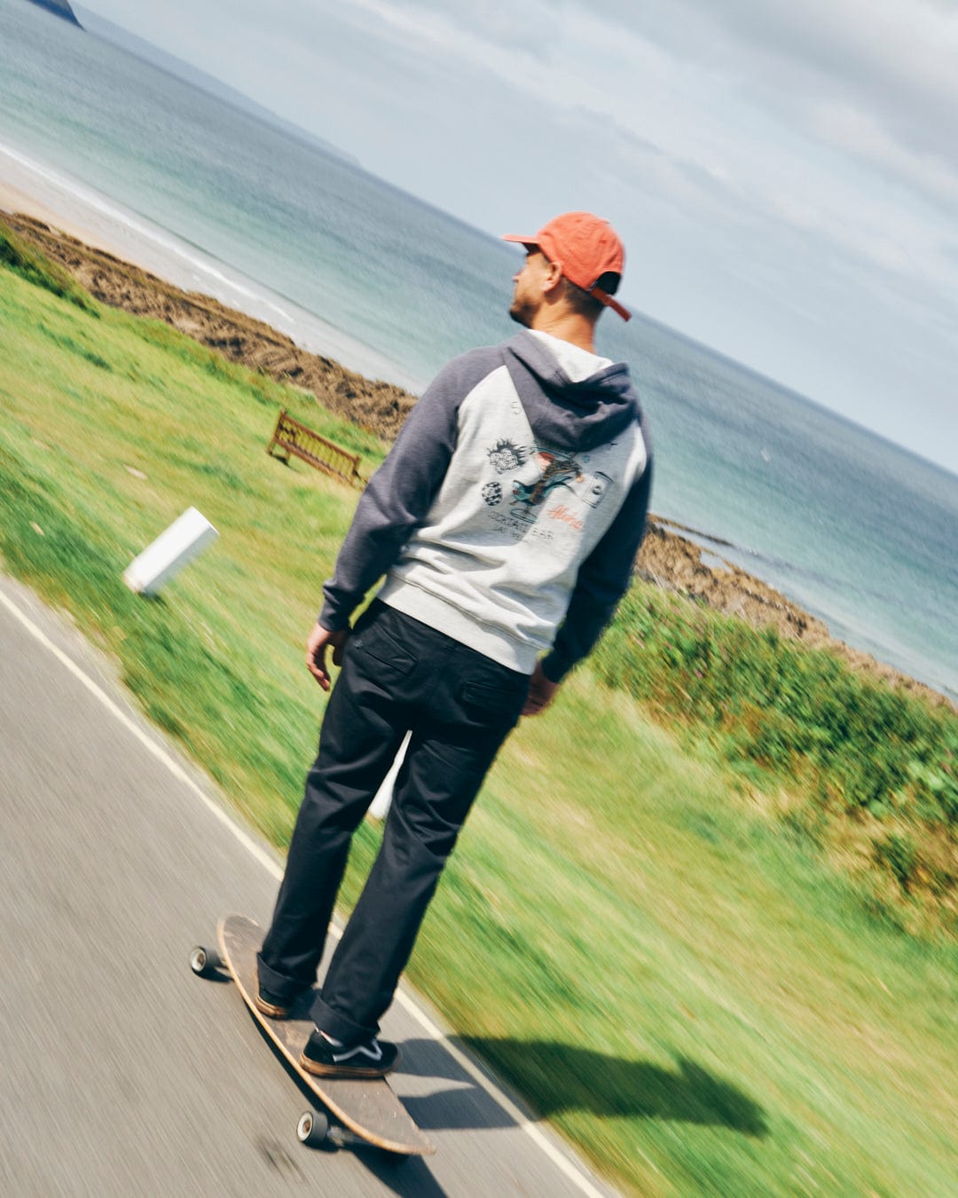 A person in a hoodie and red cap skateboards on a paved path by grassy fields, wearing Saltrock's Meltan Men's Straight Leg Twill Trouser in Dark Grey, with the ocean and sky visible in the background.