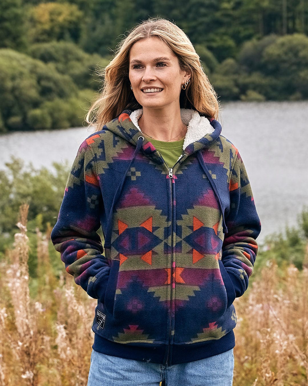 A woman wearing a Saltrock - Merida - Womens Lined Aztec Zip Fleece - Dark Blue hoodie in front of a lake.