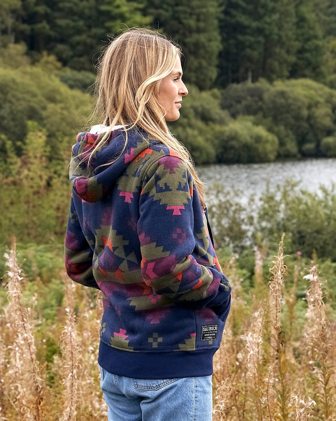 A woman wearing a Saltrock Merida - Womens Lined Aztec Zip Fleece - Dark Blue hoodie standing in a field.