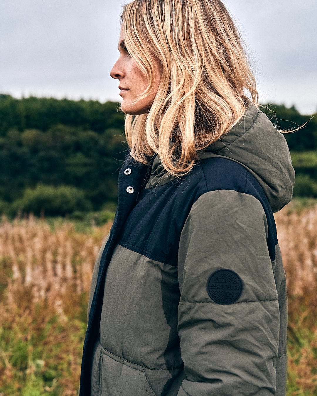 A woman is standing in a winter field wearing a Saltrock Merle - Womens Puffer Jacket - Dark Green.