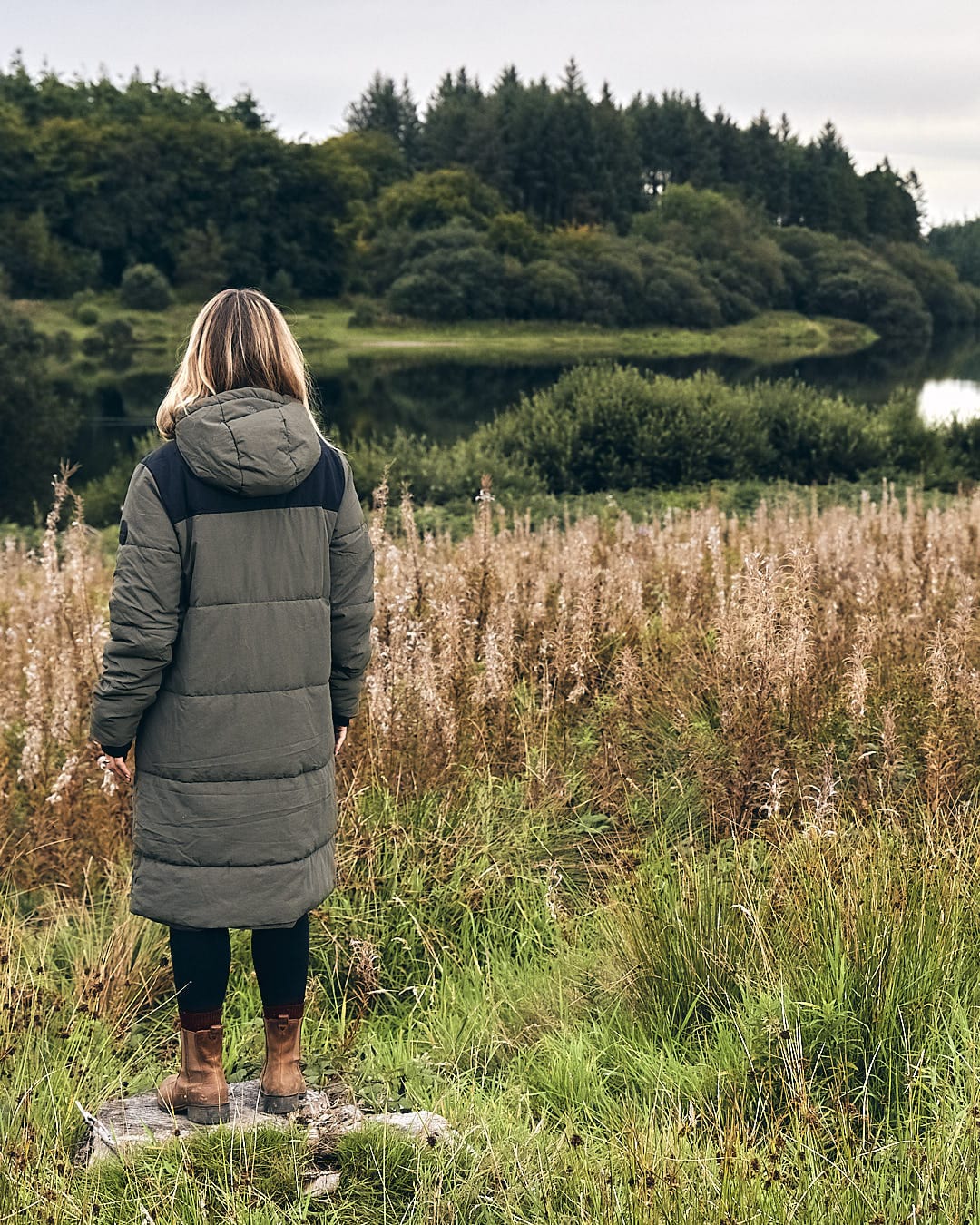 A woman in a Saltrock Merle - Womens Puffer Jacket - Dark Green is standing on a rock overlooking a lake.
