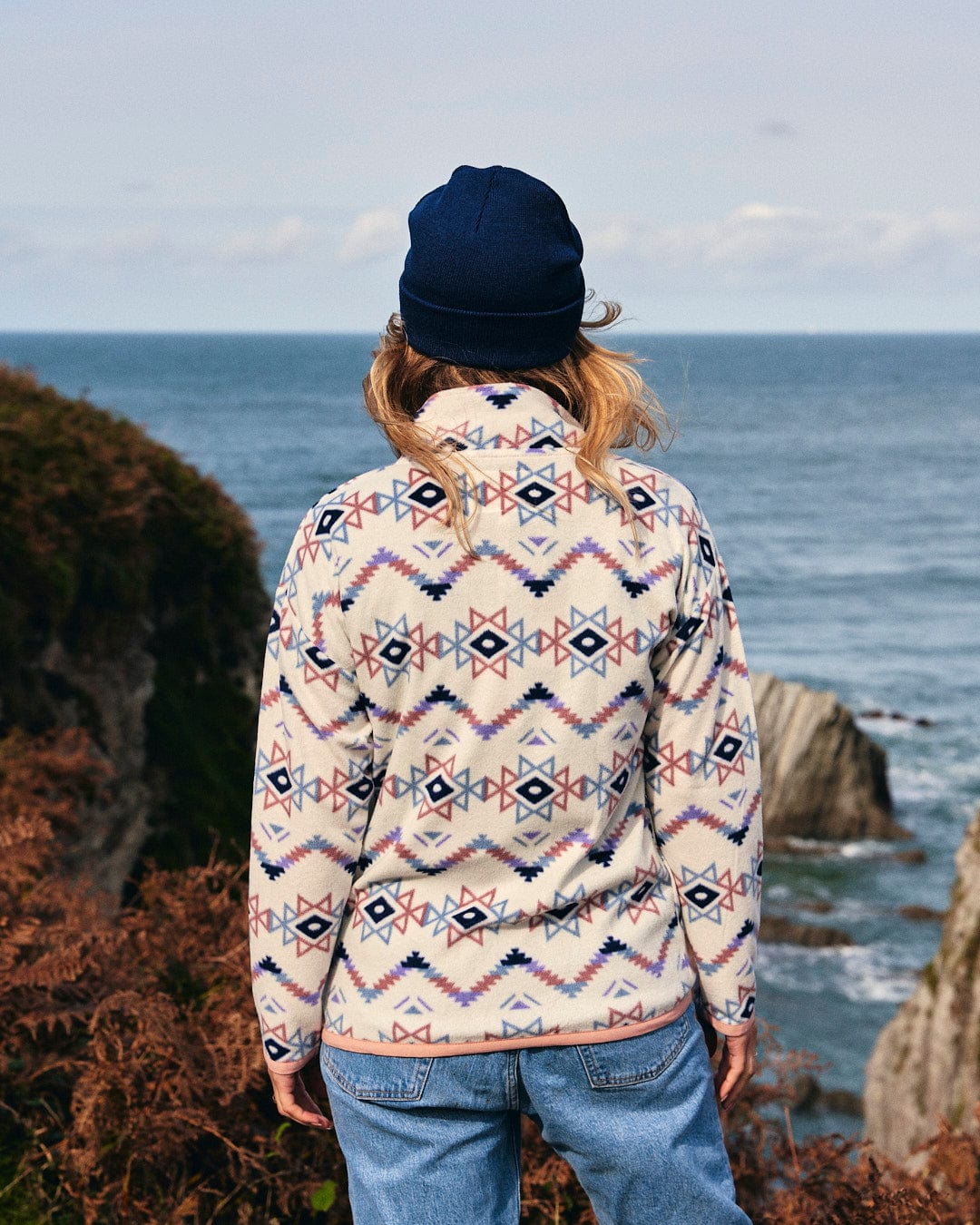 A woman is standing on a cliff overlooking the ocean wearing a Micro Ida - Womens Zip Fleece - Cream from Saltrock branding.