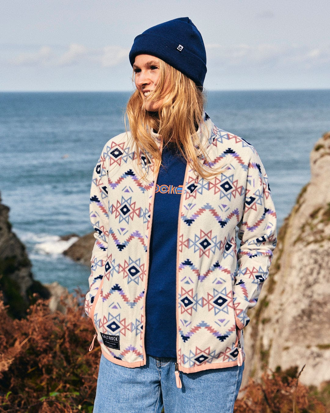 A woman wearing the Micro Ida - Womens Zip Fleece - Cream jacket standing on a rocky cliff, showcasing the Saltrock branding.