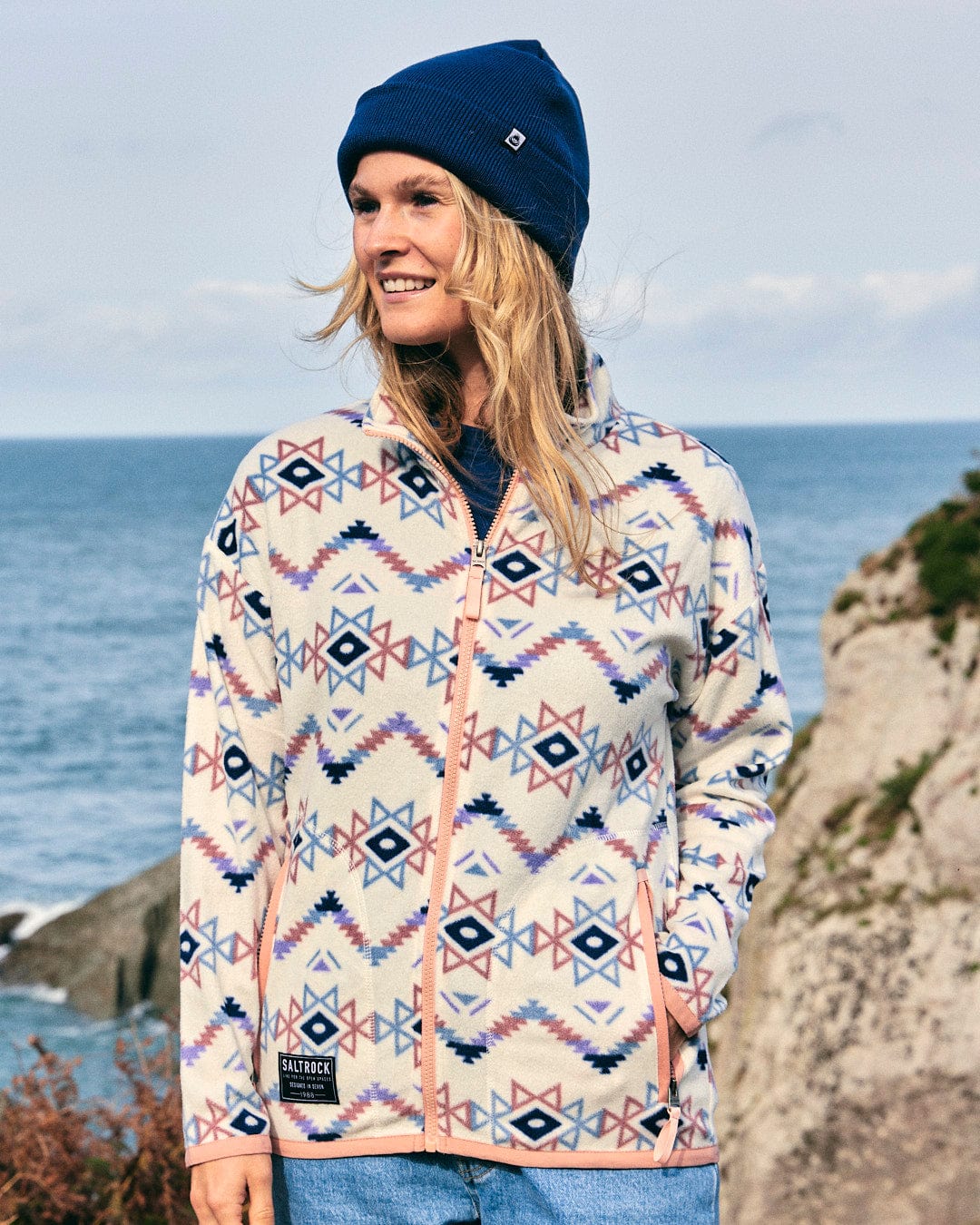 A woman wearing a Micro Ida - Womens Zip Fleece - Cream jacket, showcasing Saltrock branding, while standing by the ocean.