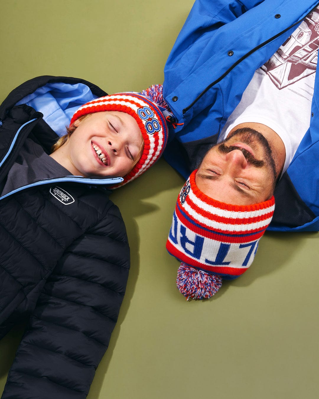 A man and a boy wearing Apres - Beanies - Blue adorned with cute pom poms lay on the ground. The beanies showcase Saltrock branding.