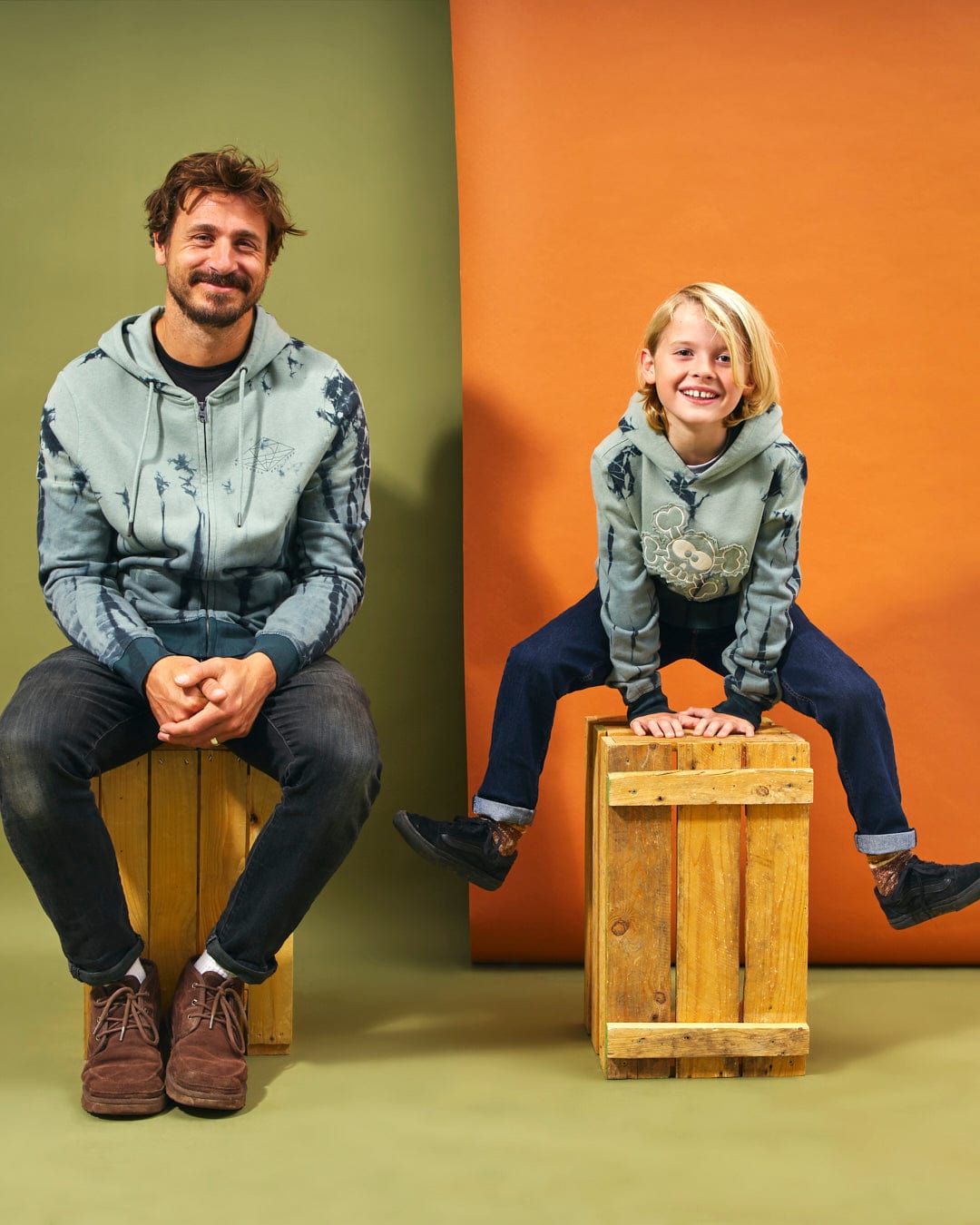 A man and a child sitting on a wooden box, both wearing tie dye print clothing - the man in a Saltrock hoodie and the child in Saltrock graphics attire.
