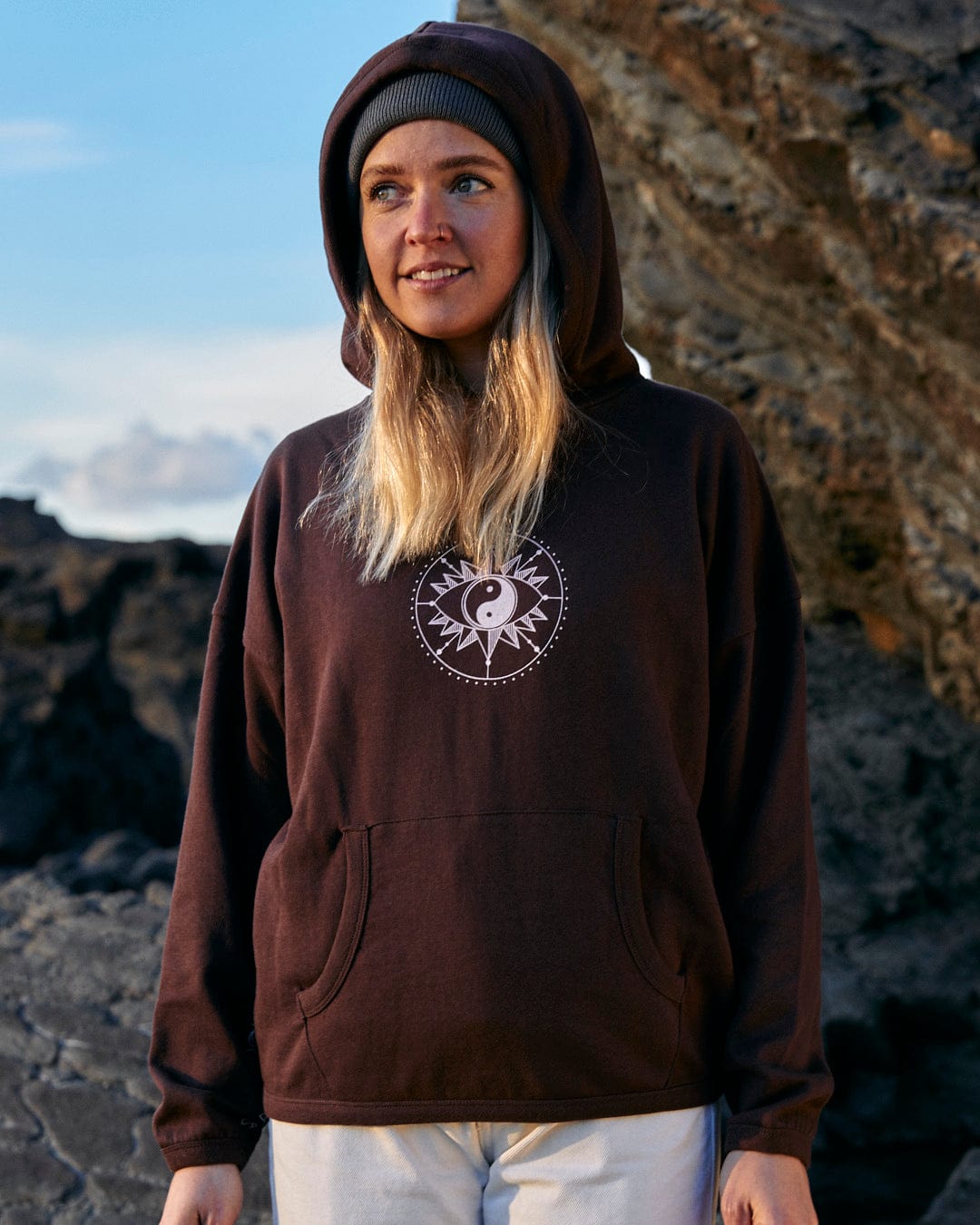 A woman wearing a Saltrock Mocha - Womens Pop Hoodie - Brown, effortlessly combining style and comfort, confidently standing on rocks.