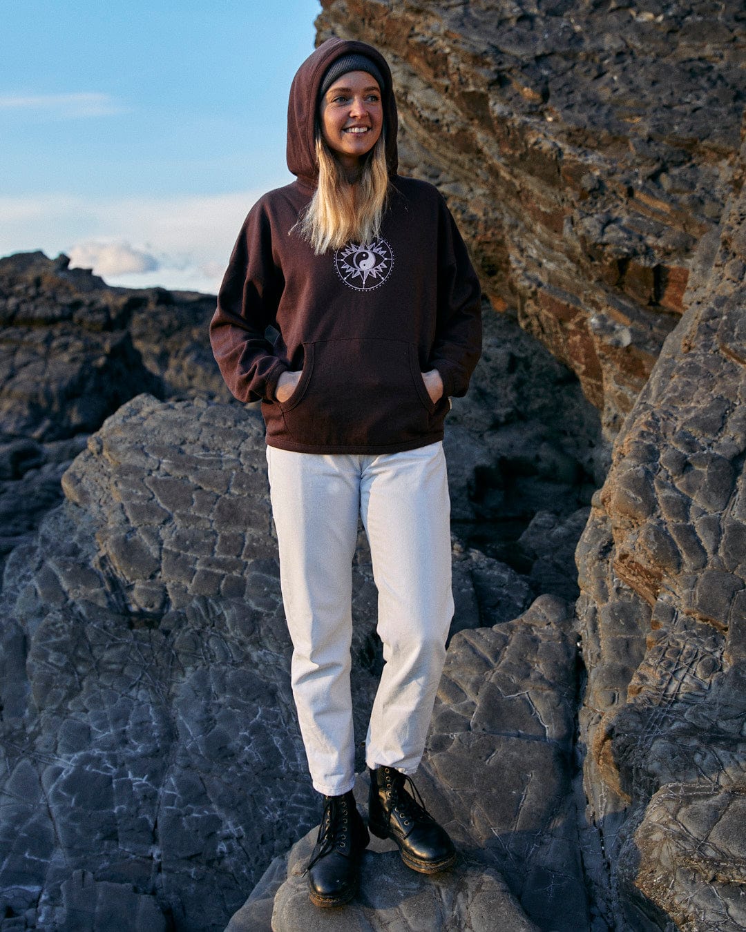 A woman showing both comfort and style by wearing a Saltrock Mocha - Womens Pop Hoodie - Brown, while standing on rocks.