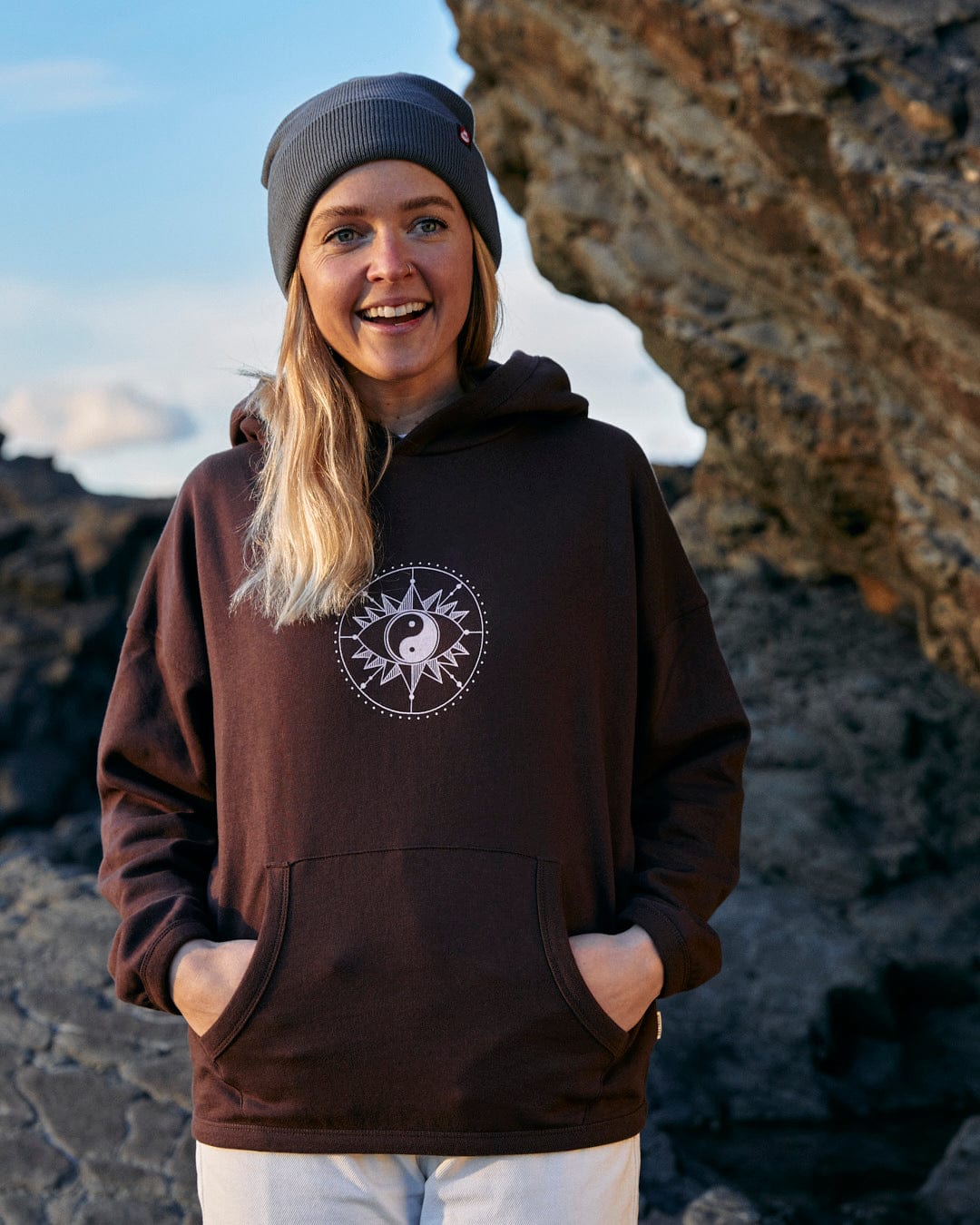 A woman wearing a Mocha - Womens Pop Hoodie - Brown, showcasing the perfect blend of style and comfort while standing on a rock, proudly displaying Saltrock branding.