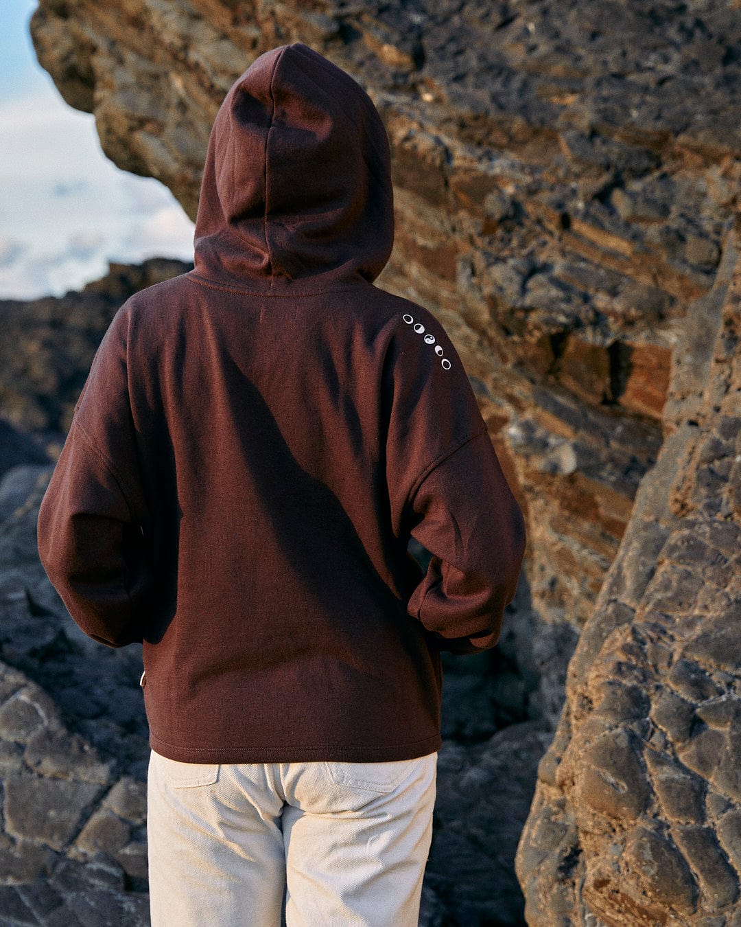 A stylish woman showcasing Saltrock branding, exuding comfort in a Mocha - Womens Pop Hoodie - Brown while standing on rocks.