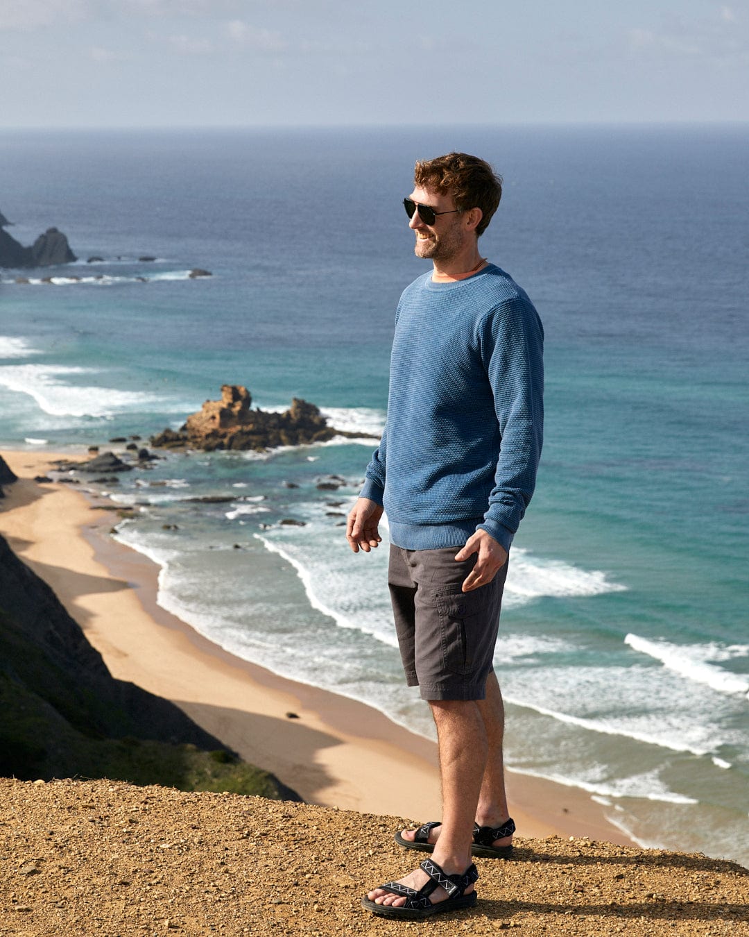 A man with a worn look standing on a cliff overlooking the ocean wearing Saltrock's Moss - Mens Washed Knitted Crew in Blue.