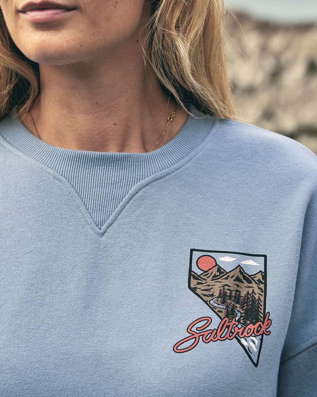 A woman wearing a Saltrock Mountain Creek Almira - Womens Balloon Sleeve Sweat - Blue sweatshirt with a mountain embroidered on it.