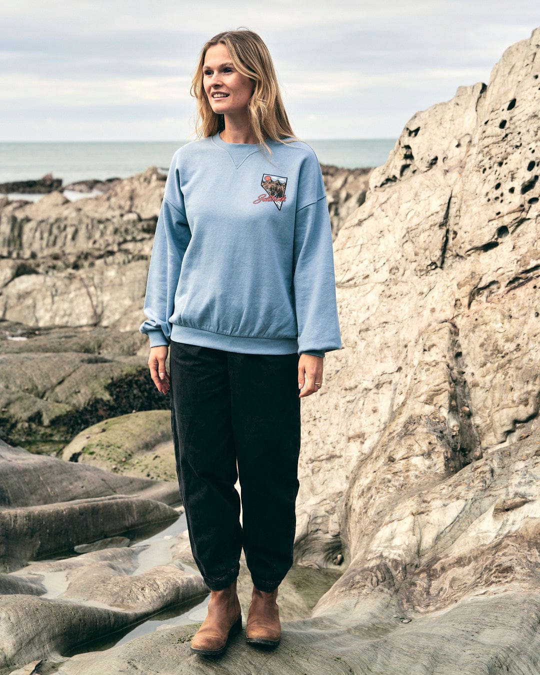 A woman wearing a Saltrock Mountain Creek Almira - Womens Balloon Sleeve Sweat - Blue sweatshirt stands on the rocks next to the ocean.