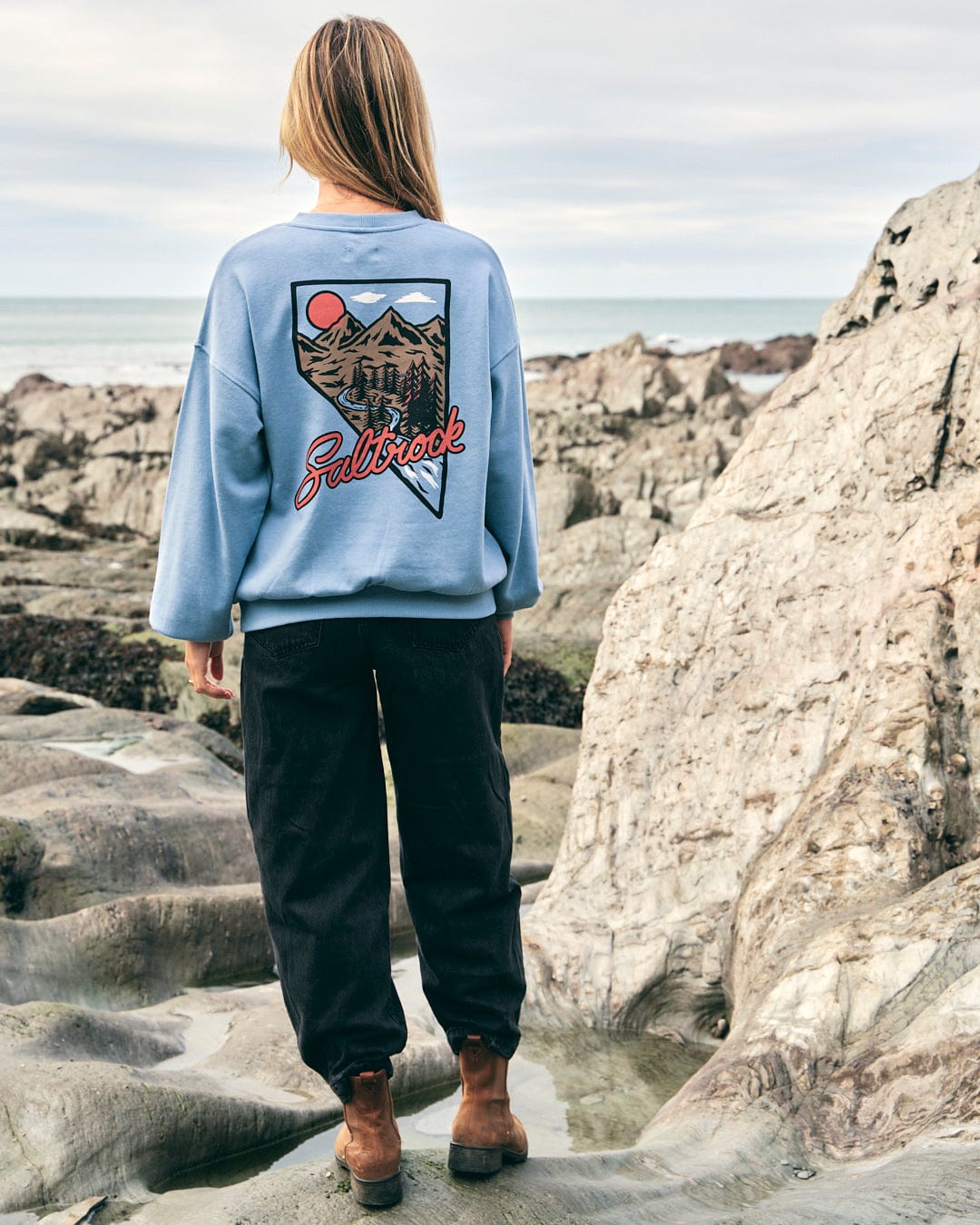 A woman wearing a Saltrock Mountain Creek Almira - Womens Balloon Sleeve Sweat - Blue sweatshirt standing on rocks near the ocean.
