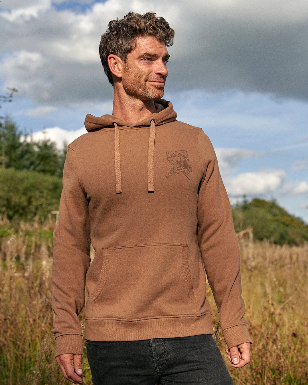 A man wearing a Saltrock Mountain Creek - Mens Pop Hoodie - Brown standing in a field with a mountain scene.