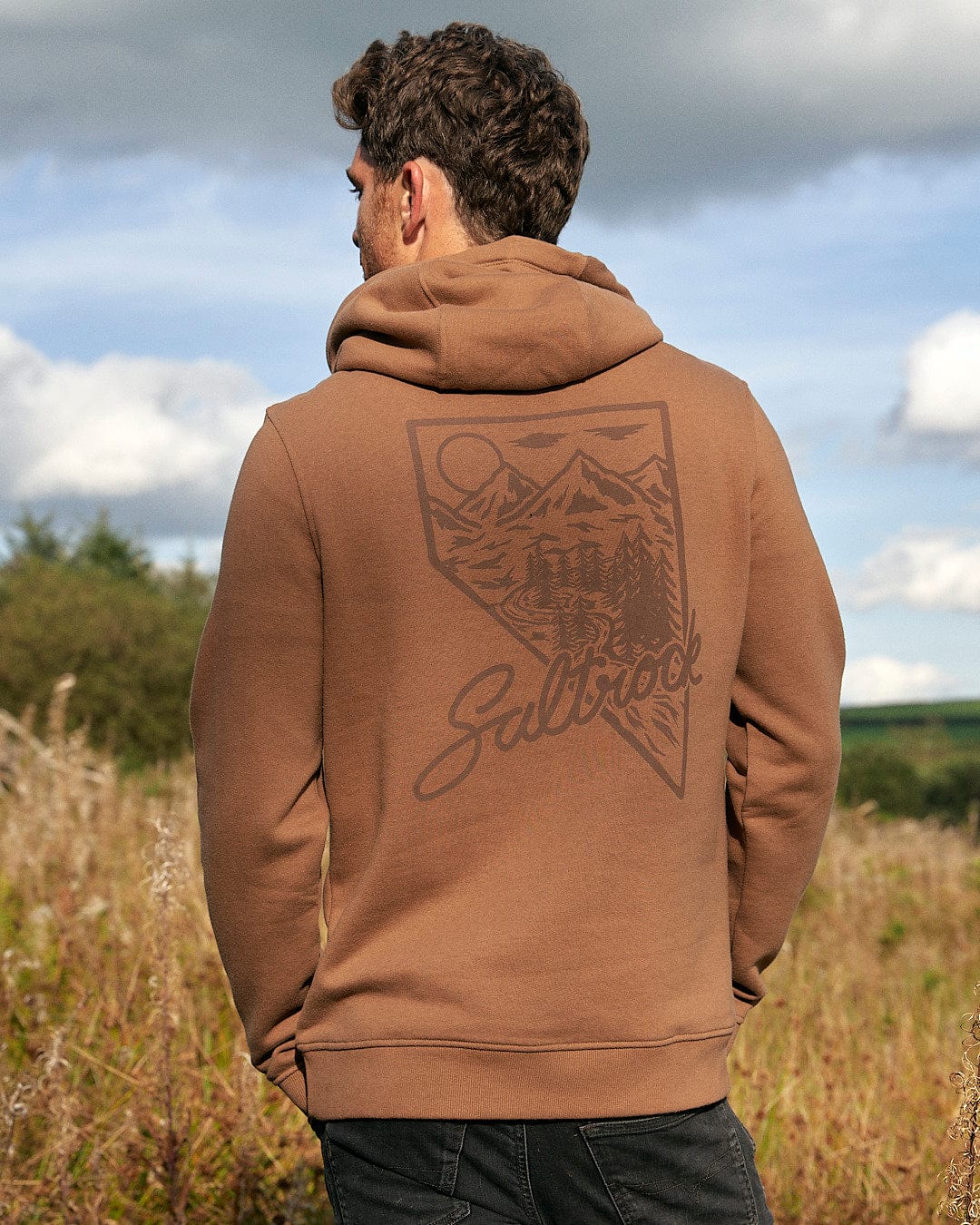 A man is standing in a mountain scene wearing a Saltrock - Mountain Creek - Mens Pop Hoodie - Brown.