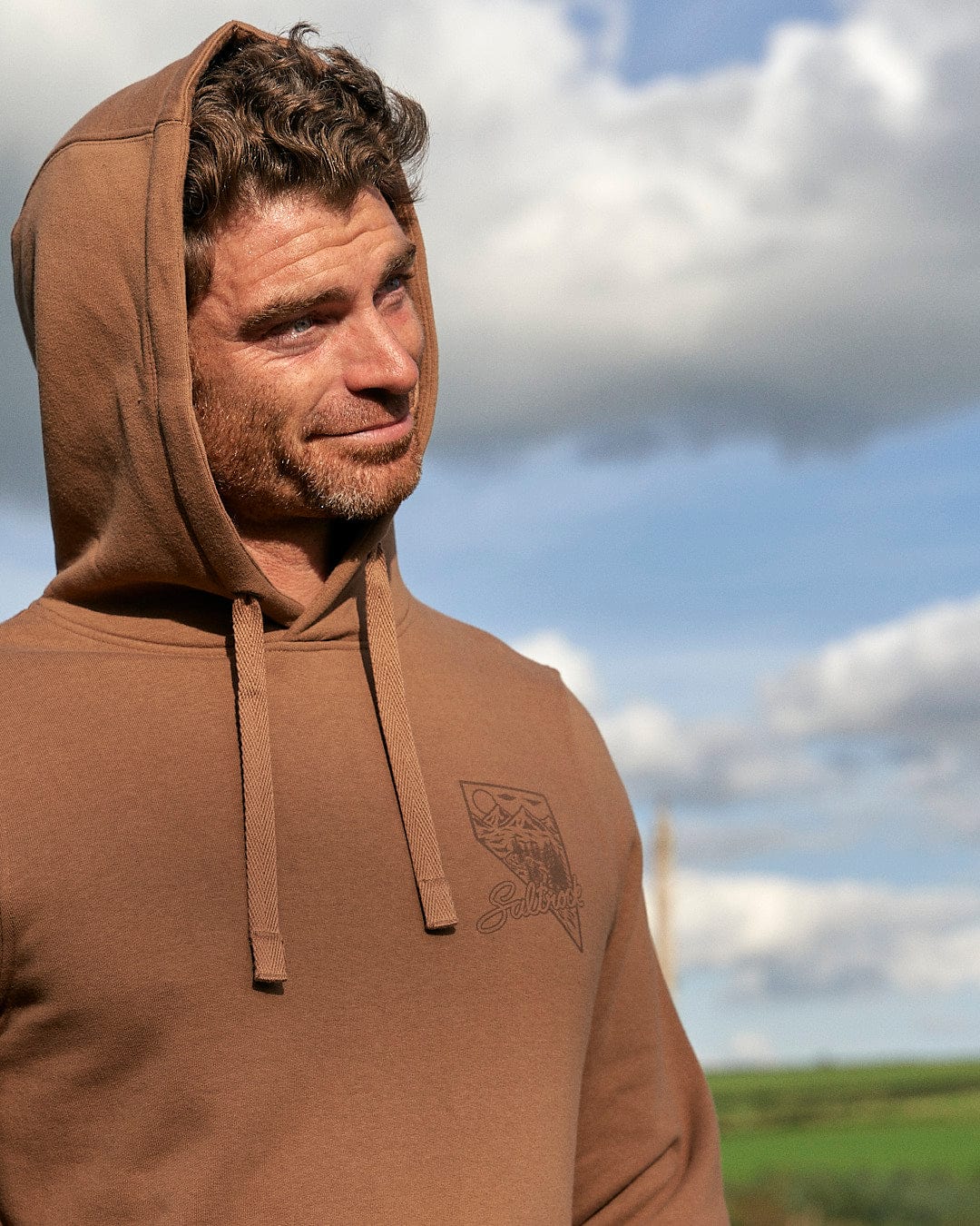 A man is standing in a mountain scene wearing a Saltrock - Mens Pop Hoodie - Brown.