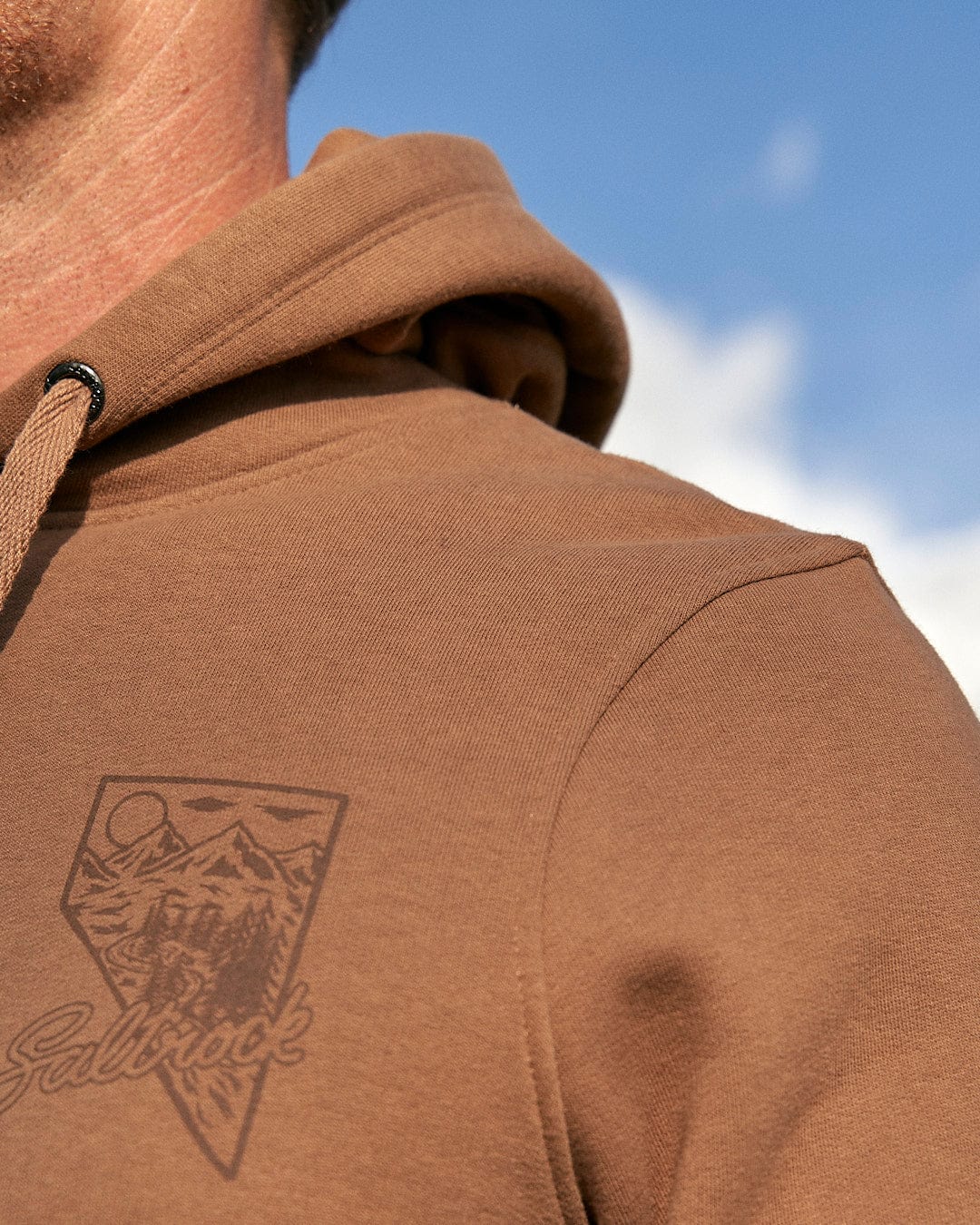 A man wearing a Saltrock Mountain Creek - Mens Pop Hoodie - Brown with a logo on it and drawstring hood.