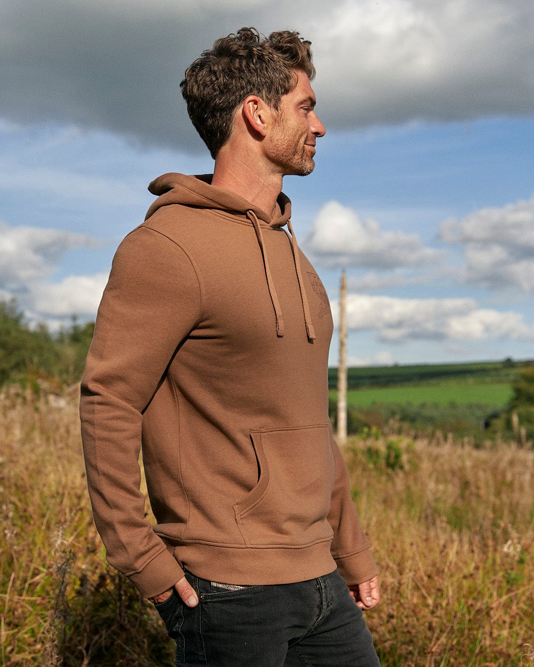 A man standing in a field wearing a Saltrock Mountain Creek - Mens Pop Hoodie - Brown with a drawstring hood.