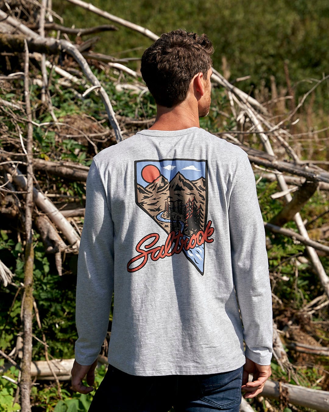 A man wearing a Saltrock Mountain Creek - Mens Long Sleeve T-Shirt - Grey standing in a wooded area.