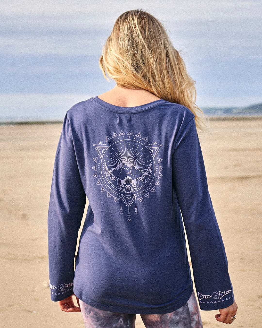 The back of a woman wearing a Saltrock - Mountain Rise Womens Flared Sleeve T-Shirt - Purple on the beach.