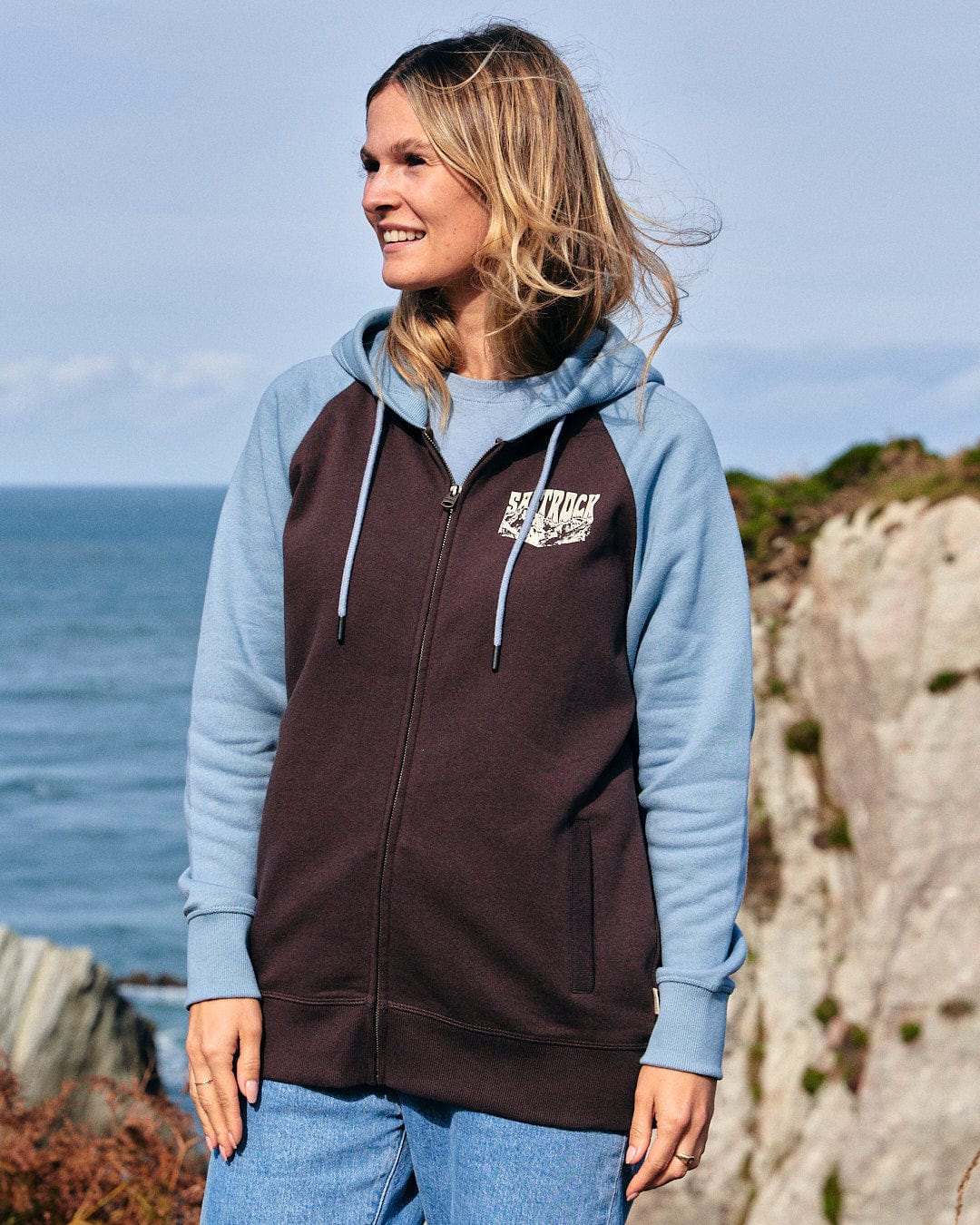 An adventurous woman wearing a brown and blue Saltrock Mountain Roaming - Womens Raglan Zip Hoodie, standing by the ocean.