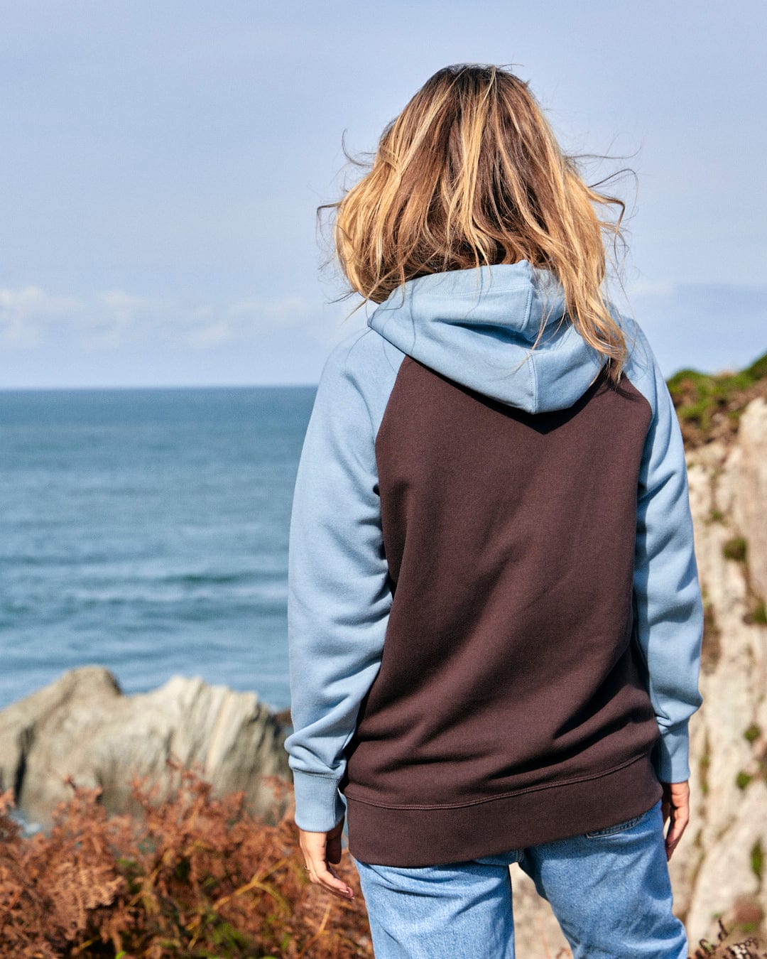 An adventurous woman, wearing a Saltrock Mountain Roaming - Womens Raglan Zip Hoodie - Brown, looking at the ocean.
