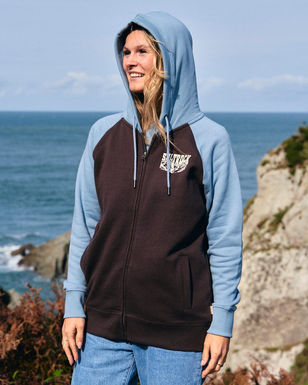 An adventurous woman wearing a Mountain Roaming - Womens Raglan Zip Hoodie by Saltrock, standing on a rock by the ocean.
